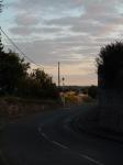    La sortie du village par un beau soir. Quelques nuages rosis par le coucher de soleil flottent dans le ciel.    